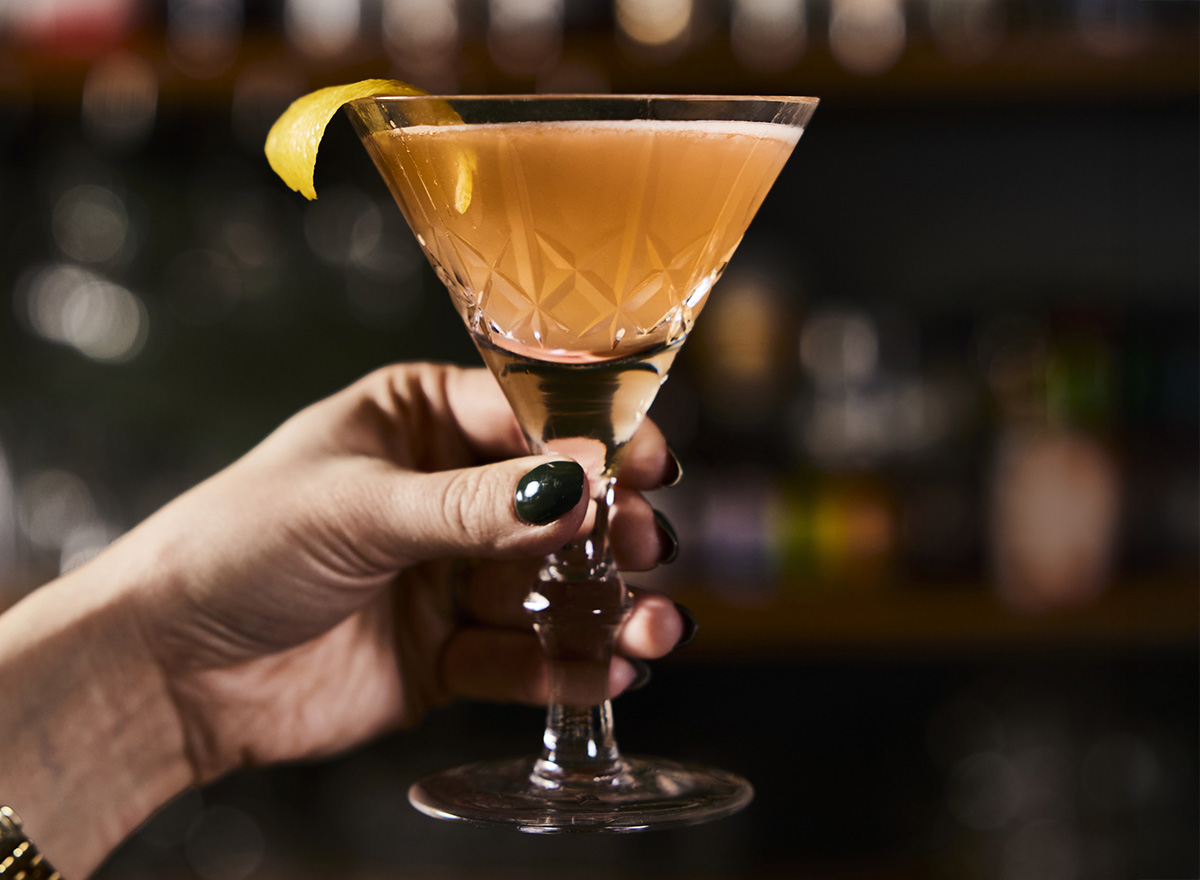 Close up of a well-manicured hand holding a cocktail glass filled with orange liquid and lemon rind garnish