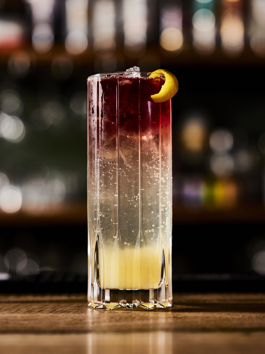 Image of a cocktail in a glass. The liquid is yellow on the bottom, clear in the middle, and red at the top, with ice and citrus rind garnish