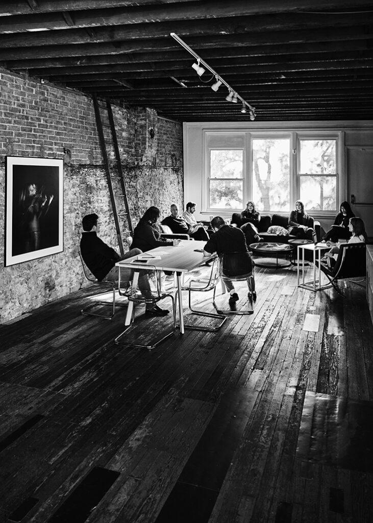 Group of coworkers sitting and holding a discussion next to large window & interior brick wall