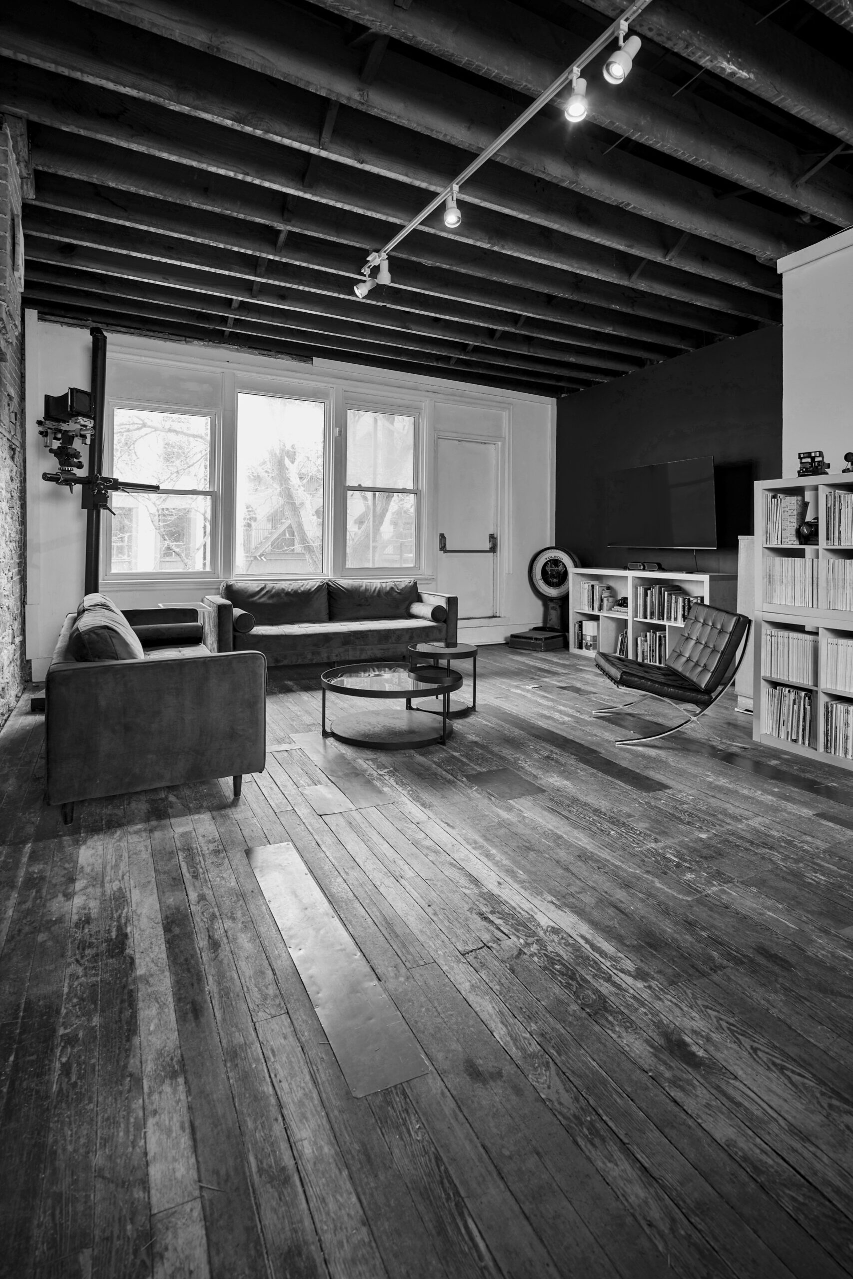 Black and white photo of view in photo studio, including 2 couches coffee table and chair against window