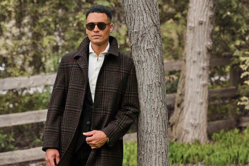 Man dressed in a suit and dress coat, wearing sunglasses and leaning against a tree